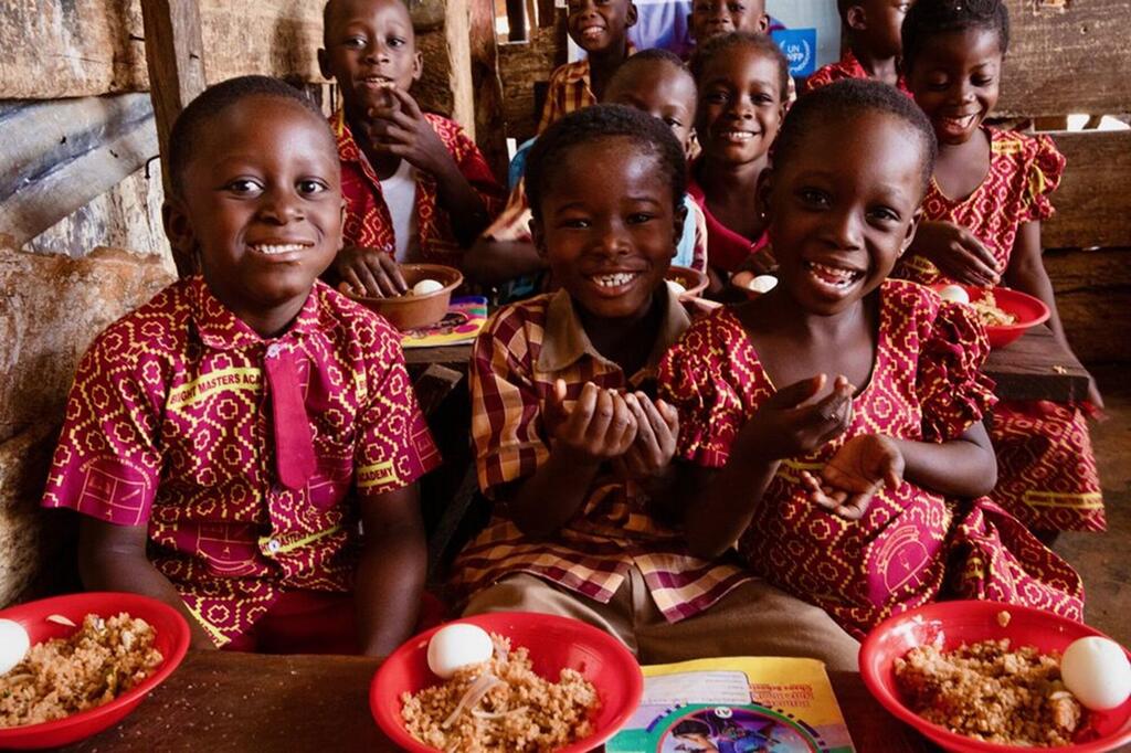 Kikundi cha watoto wa shule wa Ghana wenye tabasamu wakiwa wamevaa sare za shule wameketi mezani wakiwa na bakuli za chakula, sehemu ya programu ya chakula shuleni.