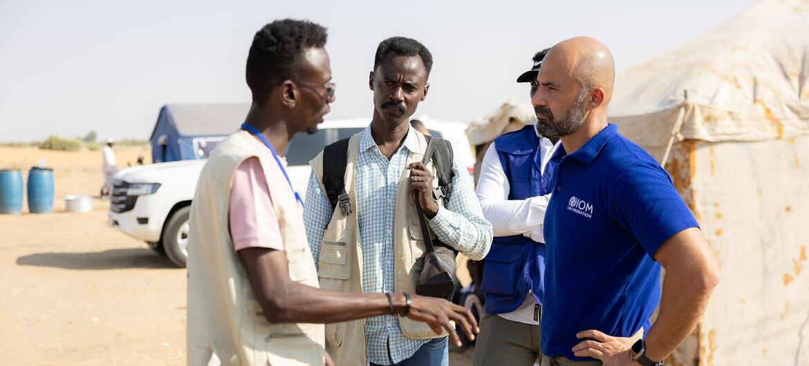 عضو من موظفي المنظمة الدولية للهجرة يرتدي سترة زرقاء يتحدث مع رجلين في موقع ميداني، مع وجود خيام ومركبات في الخلفية.
