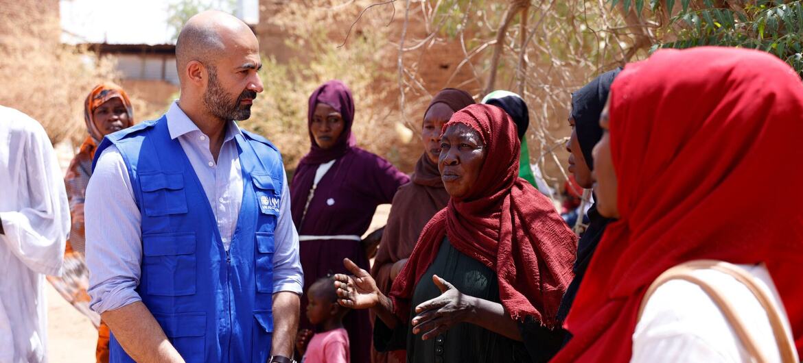 عامل إغاثة ذكر يرتدي سترة زرقاء تابعة للمنظمة الدولية للهجرة يتحدث مع مجموعة من النساء في السودان.