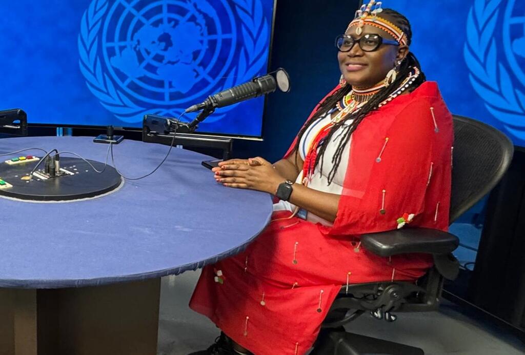 Mwanamke aliyevaa mavazi ya kitamaduni ya Kiafrika ameketi kwenye dawati akiwa na maikrofoni mbele ya skrini ya bluu inayoonyesha nembo ya Umoja wa Mataifa.