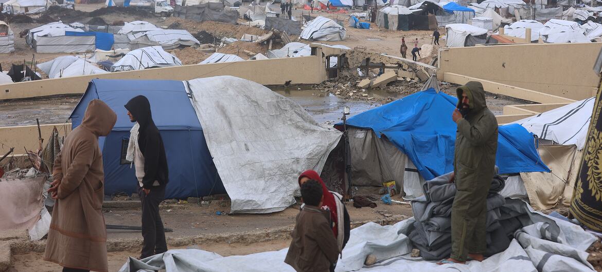 A group of people, including children, stand in a makeshift refugee camp in Gaza, surrounded by blue and white tents and debris. The background shows a vast expanse of temporary shelters near a body of water.