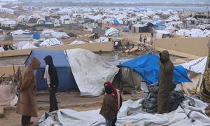 A group of people, including children, stand in a makeshift refugee camp in Gaza, surrounded by blue and white tents and debris. The background shows a vast expanse of temporary shelters near a body of water.
