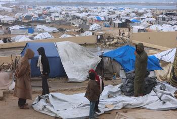 A group of people, including children, stand in a makeshift refugee camp in Gaza, surrounded by blue and white tents and debris. The background shows a vast expanse of temporary shelters near a body of water.