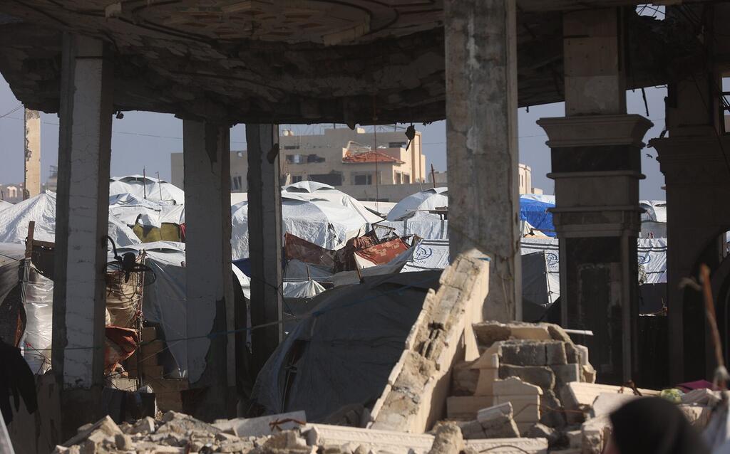 Vista a través de las ruinas de un edificio destruido en Gaza, mostrando carpas improvisadas de refugiados en el fondo bajo un cielo claro.