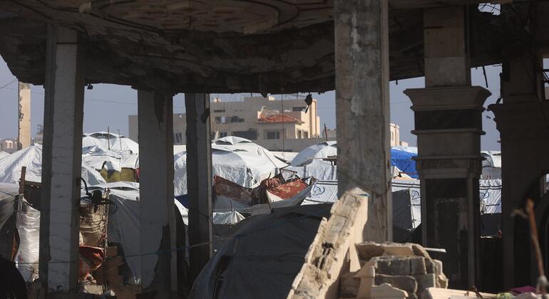 Vista a través de las ruinas de un edificio destruido en Gaza, mostrando carpas improvisadas de refugiados en el fondo bajo un cielo claro.