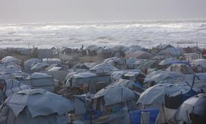 Un campamento de refugiados lleno de gente en Gaza con numerosas carpas improvisadas a lo largo de la costa, con olas que chocan en el fondo.