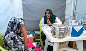 Mfanyakazi wa afya wa kike mwenye hijab ya kijani akishauriana na mwanamke wa Sudan anayeshikilia mtoto ndani ya hema la muda la matibabu nchini Sudan.