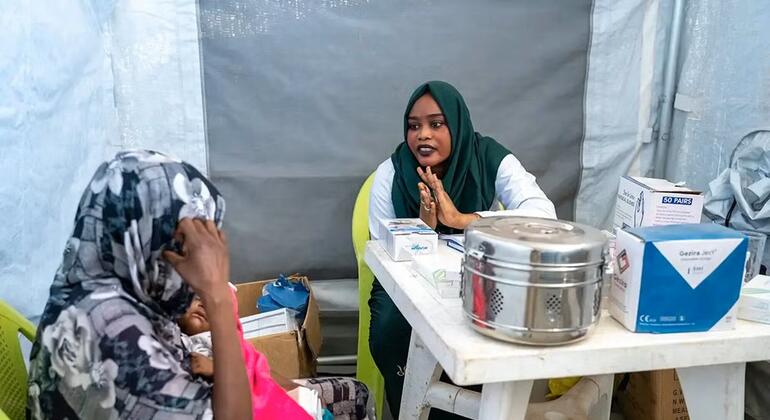 Mfanyakazi wa afya wa kike mwenye hijab ya kijani akishauriana na mwanamke wa Sudan anayeshikilia mtoto ndani ya hema la muda la matibabu nchini Sudan.