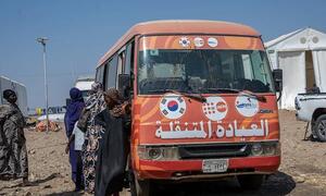 Kikundi cha wanawake wenye vifuniko vya kichwa wanasimama karibu na basi la rangi ya machungwa la kliniki ya rununu ya UNFPA katika kambi ya wakimbizi nchini Sudan. Basi lina alama za UNFPA, Korea Kusini, na mashirika mengine ya misaada, na maandishi ya Kiarabu ambayo yanasoma "Kliniki ya Rununu" mbele.