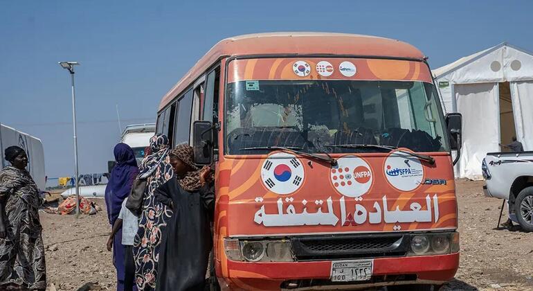 Kikundi cha wanawake wenye vifuniko vya kichwa wanasimama karibu na basi la rangi ya machungwa la kliniki ya rununu ya UNFPA katika kambi ya wakimbizi nchini Sudan. Basi lina alama za UNFPA, Korea Kusini, na mashirika mengine ya misaada, na maandishi ya Kiarabu ambayo yanasoma "Kliniki ya Rununu" mbele.