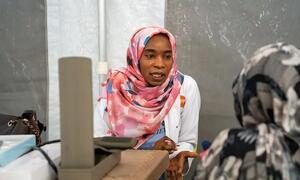 Daktari wa kike wa Sudan akiwa amevaa hijab ya rangi ya waridi na koti jeupe lenye nembo ya UNFPA akizungumza na mgonjwa katika hema la matibabu.