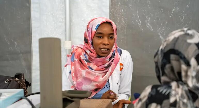 Daktari wa kike wa Sudan akiwa amevaa hijab ya rangi ya waridi na koti jeupe lenye nembo ya UNFPA akizungumza na mgonjwa katika hema la matibabu.