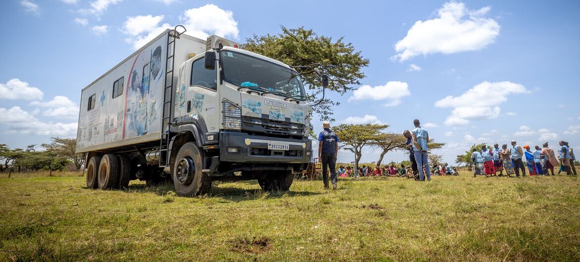 Um caminhão móvel de clínica de saúde da Organização Mundial da Saúde (OMS) estacionou num campo de erva no Quênia, com um grupo de pessoas reunidas perto, provavelmente recebendo serviços de saúde ou informações.