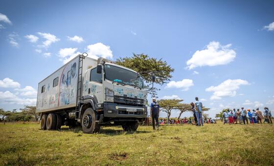 Lori la kliniki ya afya ya mkononi kutoka Shirika la Afya Ulimwenguni (WHO) lilisimama katika shamba lenye nyasi nchini Kenya, na kikundi cha watu kilikusanyika karibu, labda wakipokea huduma za afya au habari.