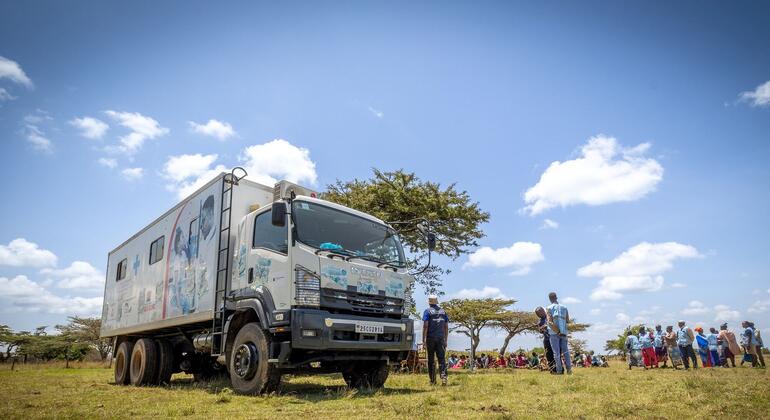 Lori la kliniki ya afya ya mkononi kutoka Shirika la Afya Ulimwenguni (WHO) lilisimama katika shamba lenye nyasi nchini Kenya, na kikundi cha watu kilikusanyika karibu, labda wakipokea huduma za afya au habari.