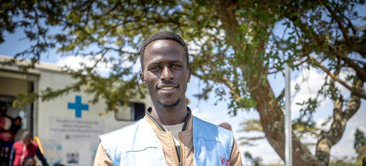 Мужской медицинский работник в жилете ВОЗ улыбается в общественной клинике в Кении, на фоне пациентов и медицинского персонала.