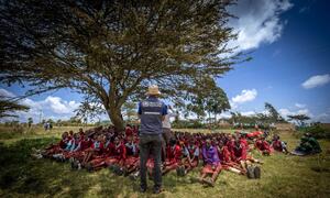 Mwanafunzi mmoja wa Shirika la Afya Ulimwenguni anazungumza na kikundi cha watoto wa shule nchini Kenya chini ya mti mkubwa, akiendeleza elimu ya afya ya uzazi.