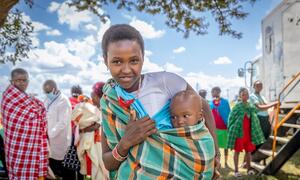 Mwanamke mwenye tabasamu kutoka Kenya aliyevalia mavazi ya Ki-Masai hubeba mtoto wake akiwa amefunikwa katika shali yenye rangi nyingi, akiwa nje na washiriki wengine wa jamii wakiwa nyuma.