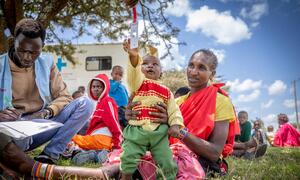 Mfanyakazi mmoja wa huduma ya afya anarekodi data huku mama mmoja wa Kenya akimshika mtoto wake, ambaye anashikilia thermometer, katika mazingira ya nje akiwa na lori la matibabu nyuma.