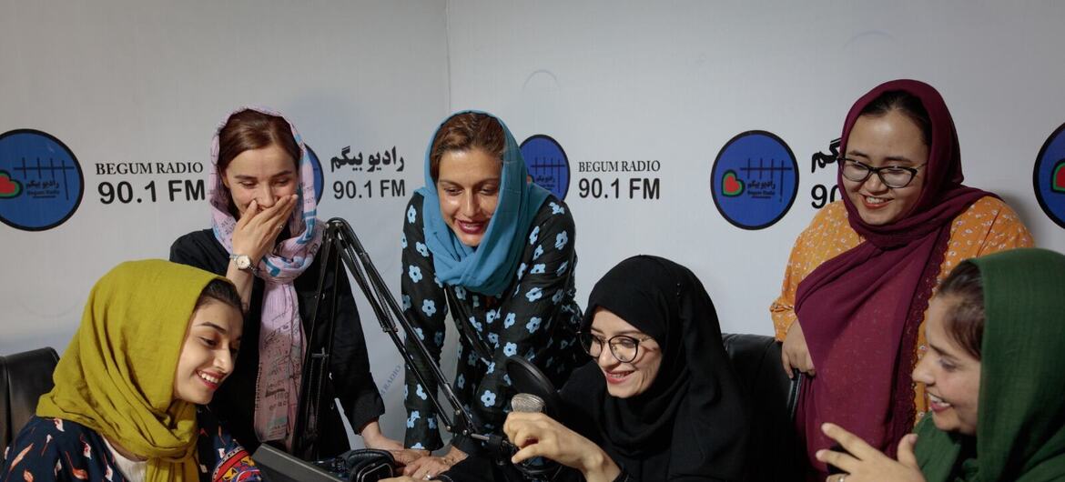 Un groupe de femmes, y compris des journalistes et des radiodiffuseurs, se rassemblent autour d'un microphone radio de Begum Radio 90.1 FM en Afghanistan, souriantes et participant à un événement de radiodiffusion.