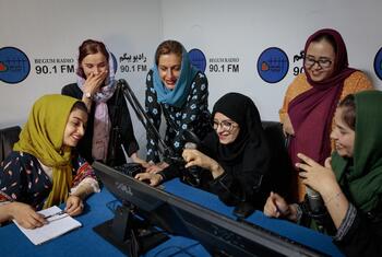 Un groupe de femmes, y compris des journalistes et des radiodiffuseurs, se rassemblent autour d'un microphone radio de Begum Radio 90.1 FM en Afghanistan, souriantes et participant à un événement de radiodiffusion.