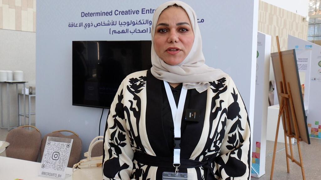 Mwanamke aliyevaa hijab na mavazi yaliyochorwa amesimama mbele ya jukwaa lililo na maandishi 'Wajasiriamali Wenye Ubunifu Walioamuliwa' katika hafla huko Manama, Bahrain, ikiendeleza uwezeshaji wa wanawake na ujasiriamali wa ubunifu.