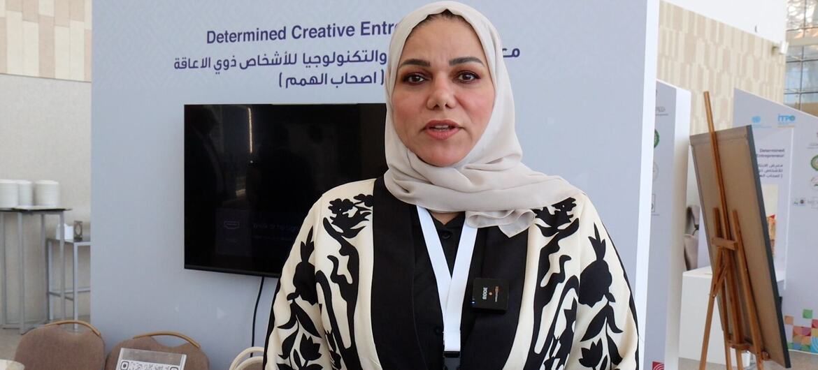 A woman wearing a hijab and a patterned outfit stands in front of a backdrop that reads 'Determined Creative Entrepreneurs' at an event in Manama, Bahrain, promoting women empowerment and creative entrepreneurship.