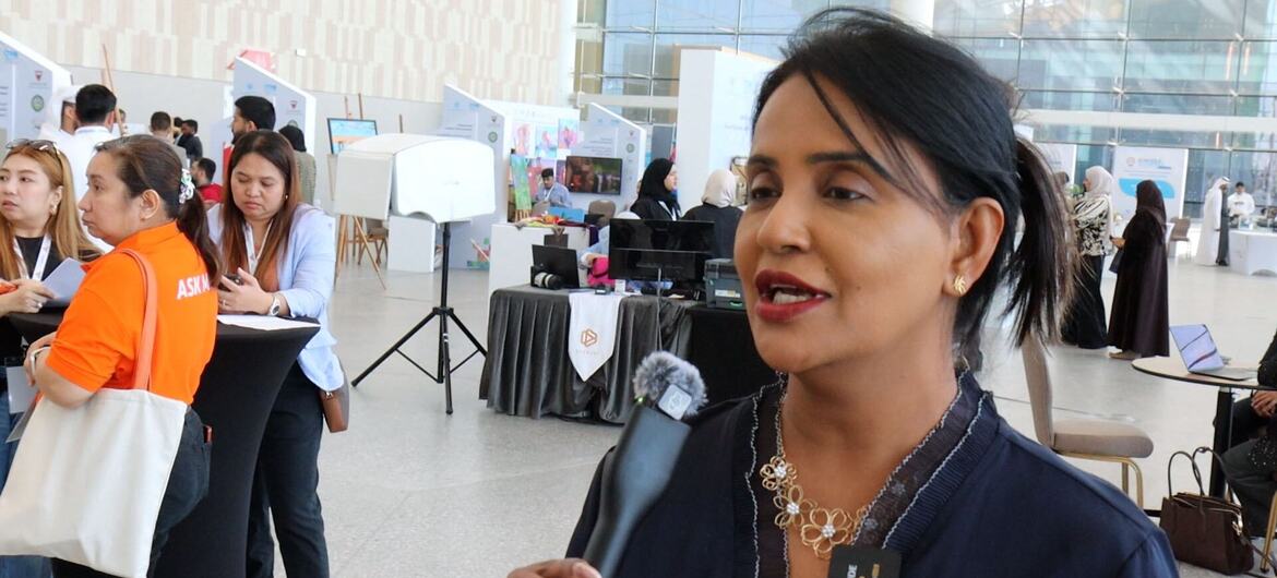 Une femme est interviewée lors d'un événement d'affaires à Manama, Bahreïn, avec d'autres participants et des stands visibles à l'arrière-plan.