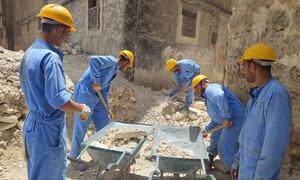 A group of construction workers in blue uniforms and yellow helmets are clearing rubble with shovels and wheelbarrows in a damaged urban area in Syria.
