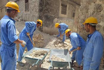 Un groupe d'ouvriers du bâtiment en uniforme bleu et casque jaune nettoie les décombres avec des pelles et des chariots dans une zone urbaine endommagée en Syrie.