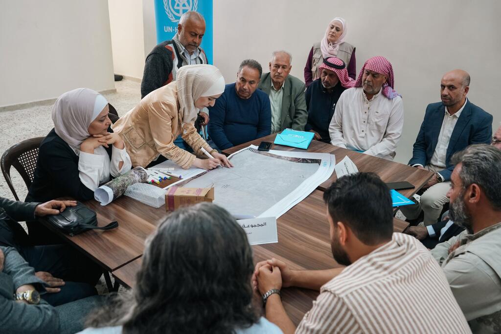 Un groupe de personnes, y compris des hommes et des femmes portant des vêtements traditionnels et des foulards, sont rassemblés autour d'une table dans un cadre d'atelier, révisant et discutant de grands plans architecturaux. Une bannière de l'ONU-Habitat est visible à l'arrière-plan. L'atelier fait partie d'un effort participatif visant à établir des priorités de relance pour le logement en Syrie, financé par plusieurs agences des Nations Unies.