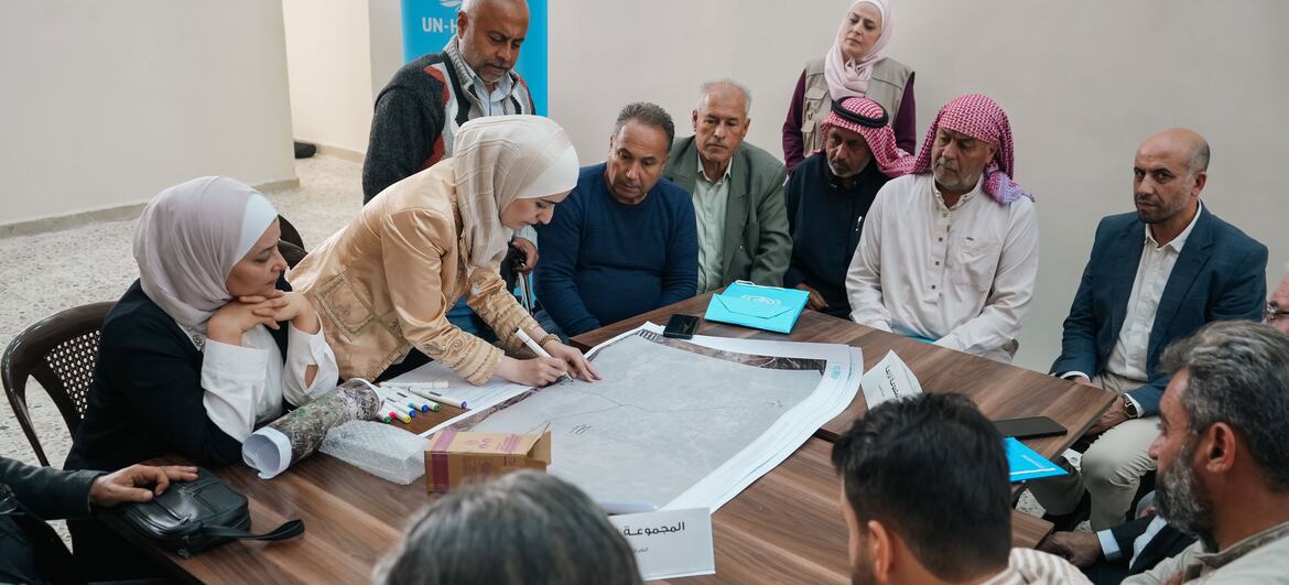 A group of people, including men and women wearing traditional attire and headscarves, are gathered around a table in a workshop setting, reviewing and discussing large architectural plans. A UN-Habitat banner is visible in the background. The workshop is part of a participatory effort to set recovery priorities for housing in Syria, funded by multiple UN agencies.
