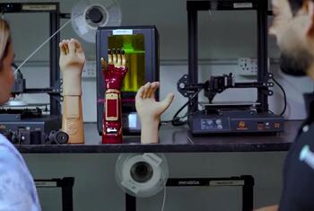 Two individuals observing AI-powered prosthetic limbs displayed on a workbench with 3D printers in the background.
