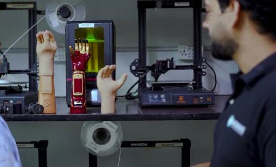 دو افراد اے آئی سے چلنے والے مصنوعی اعضاء کا مشاہدہ کر رہے ہیں جو پس منظر میں تھری ڈی پرنٹرز کے ساتھ ورک بینچ پر دکھائے گئے ہیں۔
