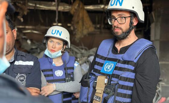 Olga Cherevko, UNOCHA spokesperson, standing in a damaged building in Gaza alongside other UN staff wearing blue vests and helmets.