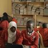 Wanafunzi wawili wa Kenya wenye sare nyekundu za shule, msichana mmoja amevaa hijab nyeupe, wanafanya kazi pamoja kwenye kompyuta ndogo katika maktaba ya shule iliyojaa karatasi.