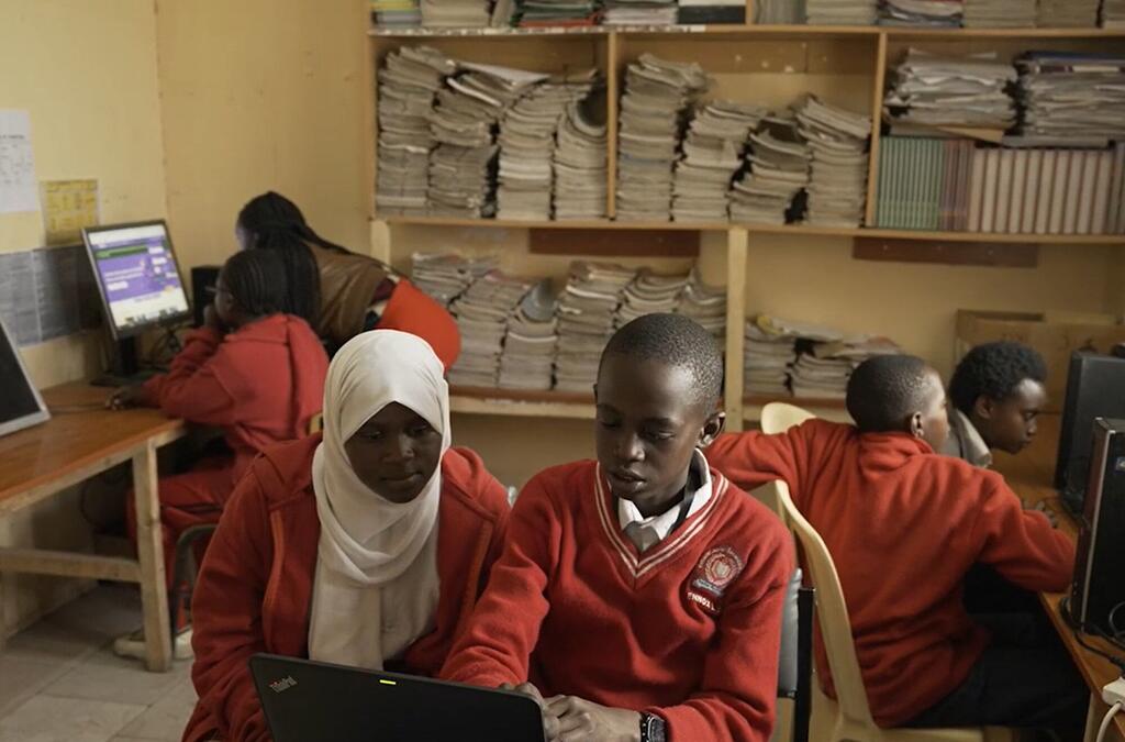 Wanafunzi wawili wa Kenya wenye sare nyekundu za shule, msichana mmoja amevaa hijab nyeupe, wanafanya kazi pamoja kwenye kompyuta ndogo katika maktaba ya shule iliyojaa karatasi.