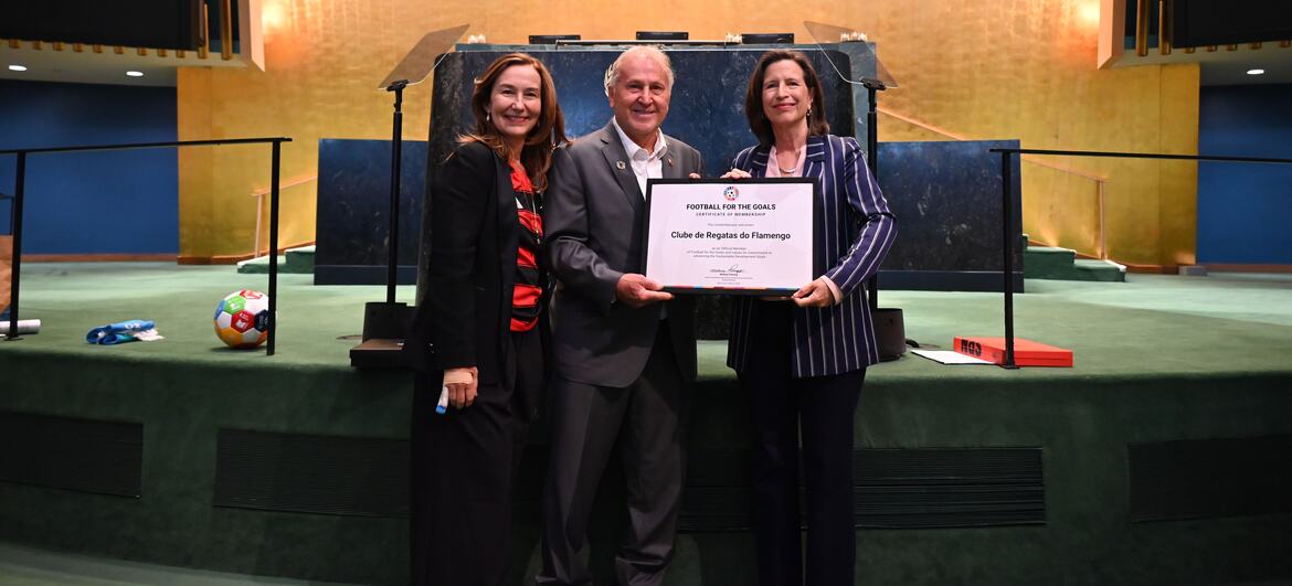 Melissa Fleming, Subsecretária-Geral da ONU para as Comunicações Globais, e uma mulher com um blazer listrado apresentam um certificado do Football for Goals à lenda do futebol brasileiro Zico (Arthur Antunes Coimbra) em um palco das Nações Unidas.