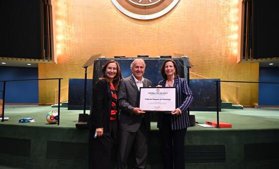 Melissa Fleming, Subsecretária-Geral da ONU para as Comunicações Globais, e uma mulher com um blazer listrado apresentam um certificado do Football for Goals à lenda do futebol brasileiro Zico (Arthur Antunes Coimbra) em um palco das Nações Unidas.