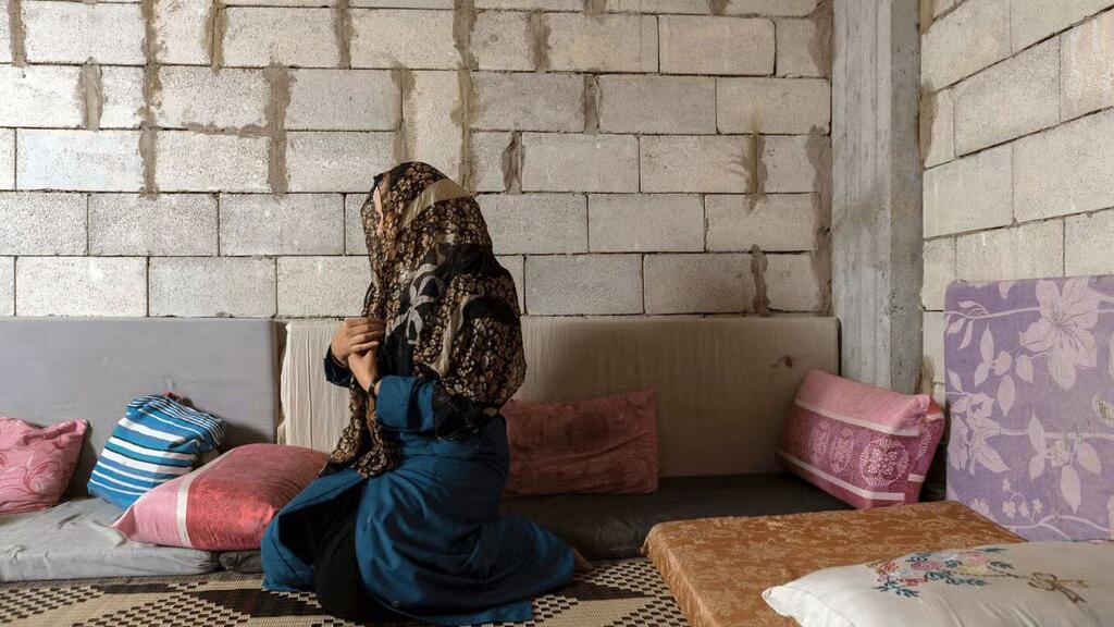 Une femme syrienne vêtue d'un foulard et d'une robe bleue s'agenouille en prière à l'intérieur d'un simple abri aux murs en parpaings.