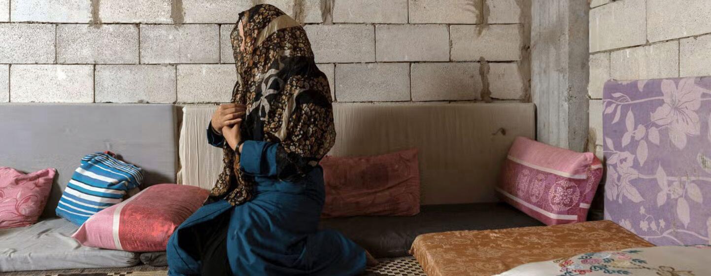 A Syrian woman in a headscarf and blue dress kneels in prayer inside a simple shelter with cinder block walls.