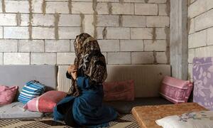 A Syrian woman in a headscarf and blue dress kneels in prayer inside a simple shelter with cinder block walls.