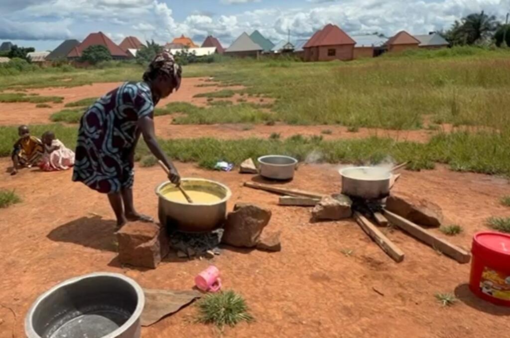 Mwanamke anapika sufuria kubwa ya chakula juu ya moto wa wazi katika kijiji cha Tanzania, na watoto wameketi karibu kama sehemu ya mpango wa FAO wa msaada wa lishe.
