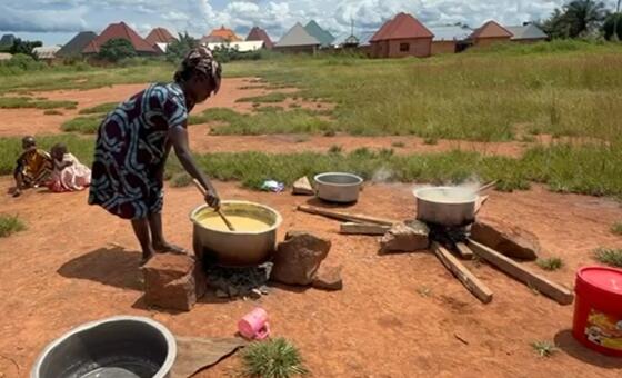Mwanamke anapika sufuria kubwa ya chakula juu ya moto wa wazi katika kijiji cha Tanzania, na watoto wameketi karibu kama sehemu ya mpango wa FAO wa msaada wa lishe.
