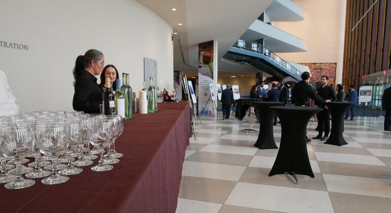 Uma área de recepção para uma celebração do Dia da Língua Portuguesa, com uma mesa longa contendo garrafas de vinho e copos, enquanto os participantes fazem networking ao fundo.