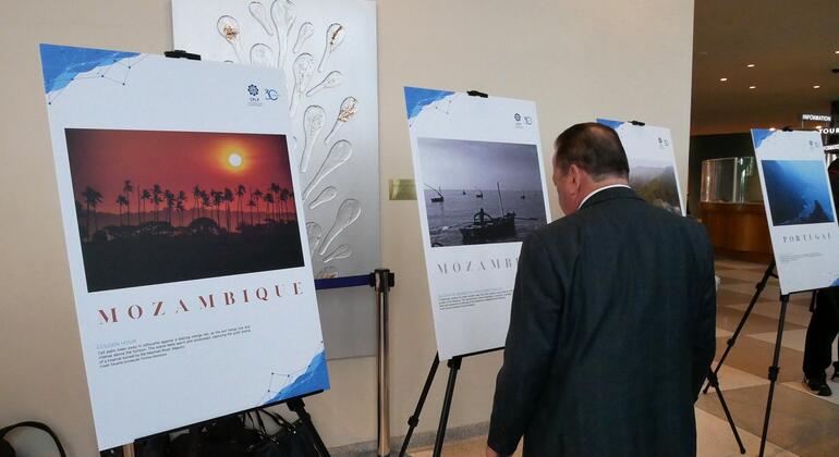 Um homem de terno observa cartazes em uma exposição que celebra o Dia da Língua Portuguesa, com um cartaz apresentando um pôr do sol em Moçambique.