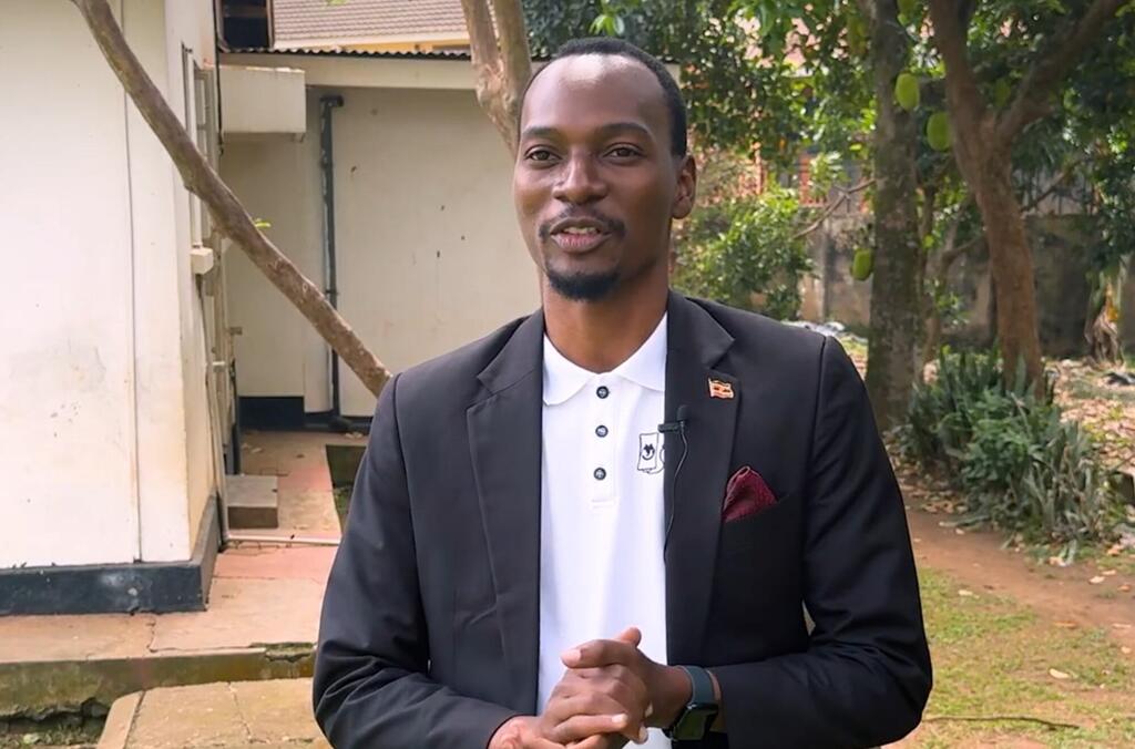 Mfanyakazi mmoja wa afya nchini Uganda, akiwa amevaa blazer nyeusi na shati nyeupe ya polo na pini ya bendera ya Uganda, anasimama nje mbele ya jengo lenye miti kwenye mandhari.