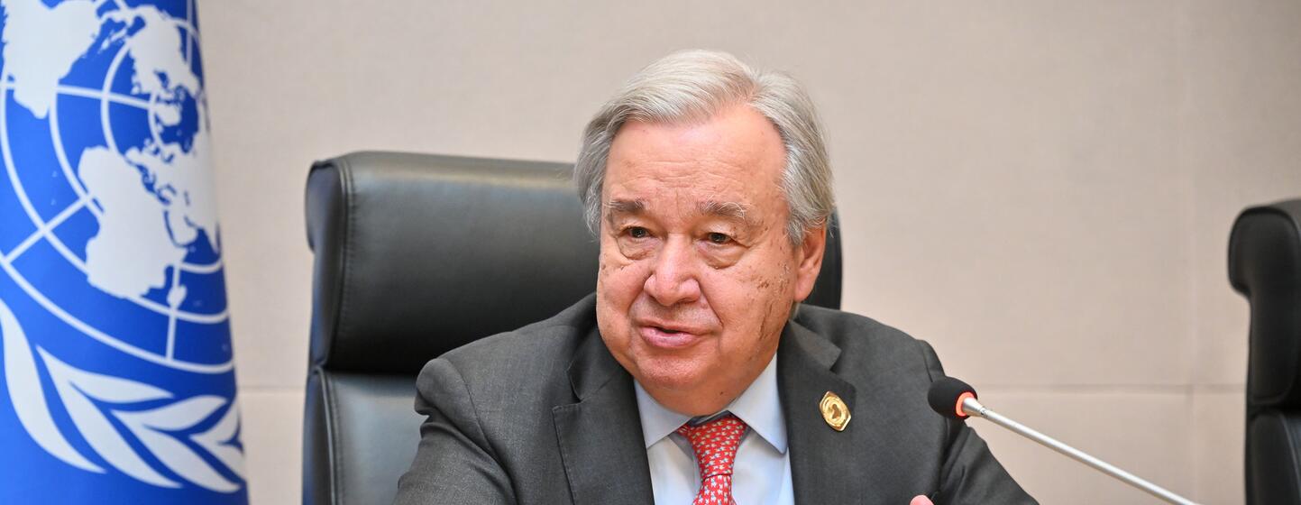 António Guterres, secrétaire général des Nations unies, assis à une table de conférence avec un micro et le drapeau de l'ONU en arrière-plan.