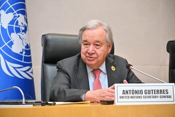 António Guterres, the United Nations Secretary-General, seated at a conference table with a microphone and UN flag in the background.