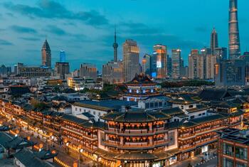 A panoramic view of Shanghai's skyline at dusk, featuring modern skyscrapers like the Shanghai Tower and Oriental Pearl Tower in the background, and traditional Chinese architecture with lit-up buildings in the foreground.